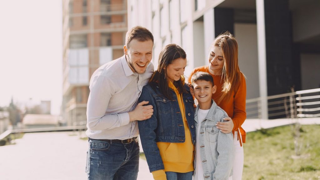 Delighted family in trendy casual wear holding each other and standing near modern building on city street while walking together during weekend